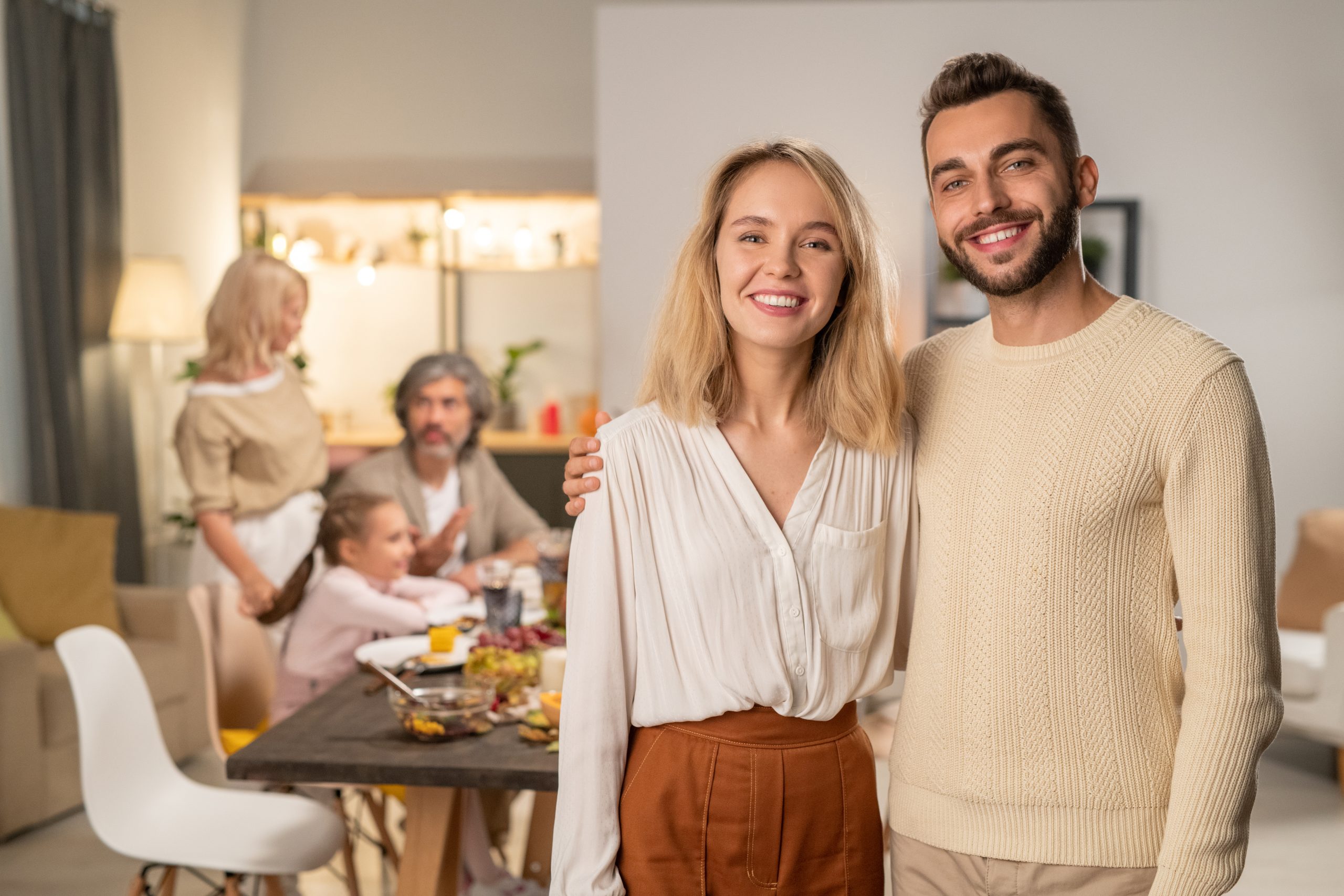 Happy young affectionate couple in casualwear looking at you with toothy smiles on background of their parents and children by festive table