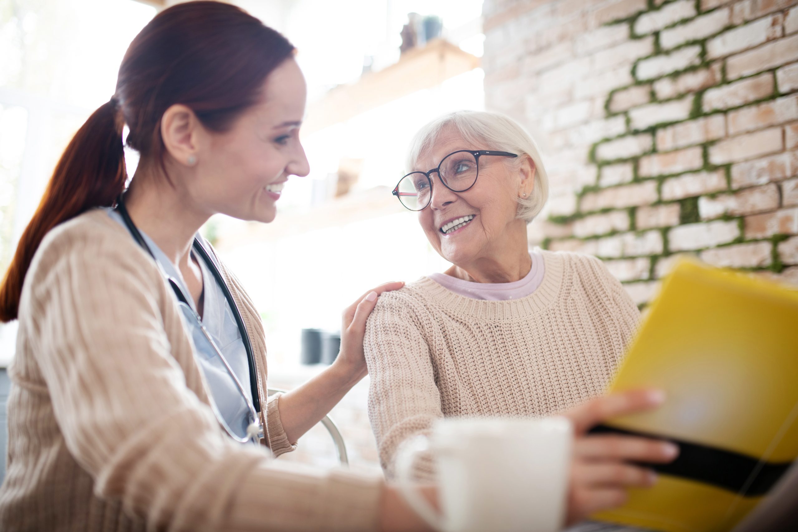 Kind aged woman. Red-haired nurse smiling while talking to kind aged woman reading the book